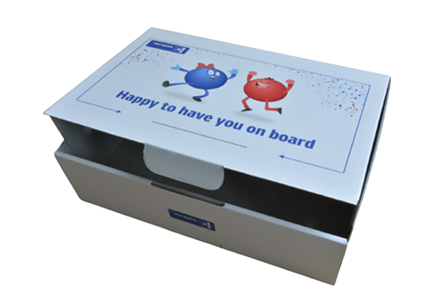 Pudełko z napisem „Cieszymy się, że jesteś z nami” na wierzchu, zdobione kolorowymi postaciami: niebieską i czerwoną, które tańczą i wyrażają radość.