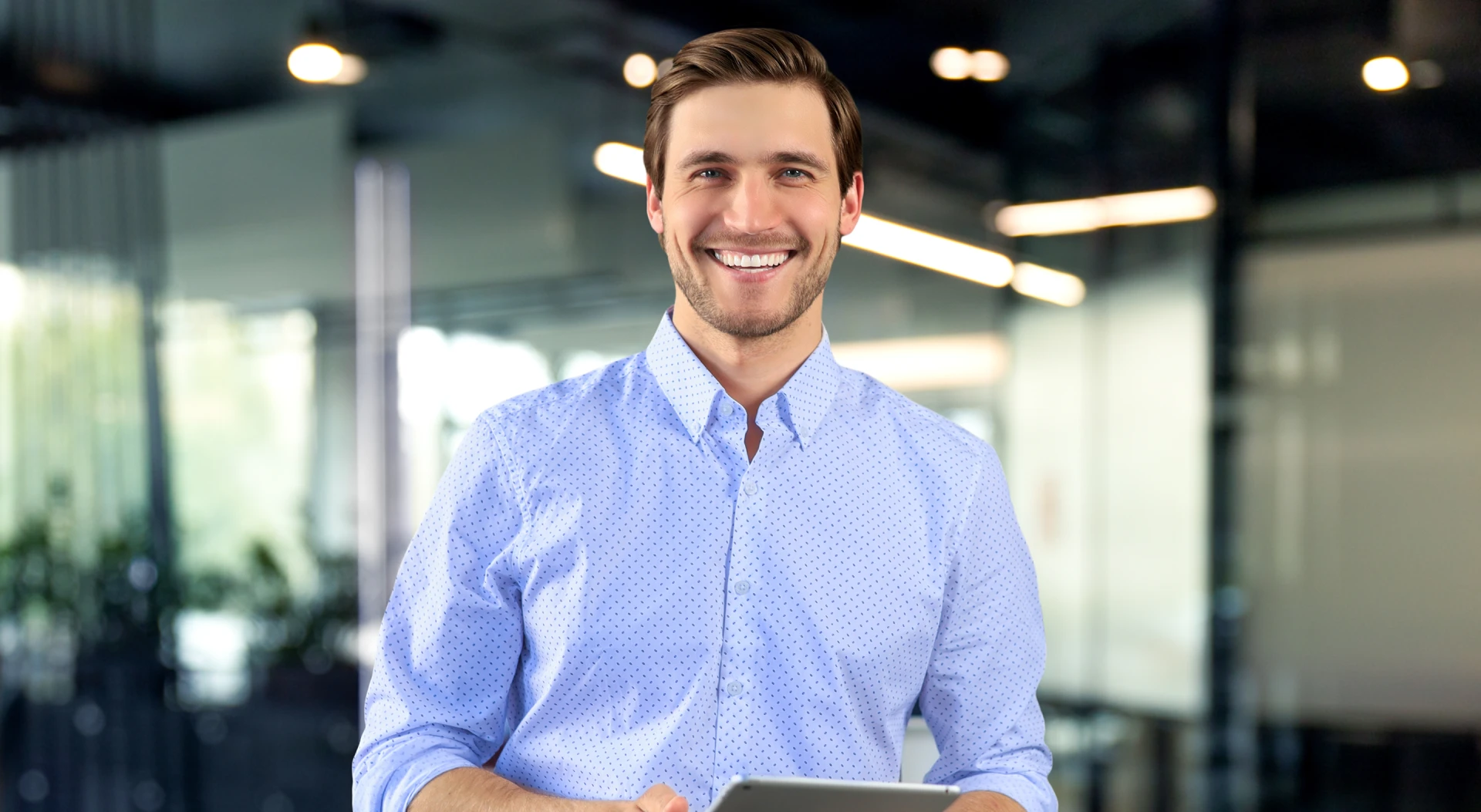 Mężczyzna w jasnej koszuli uśmiecha się, trzymając tablet. W tle widać nowoczesne biuro.