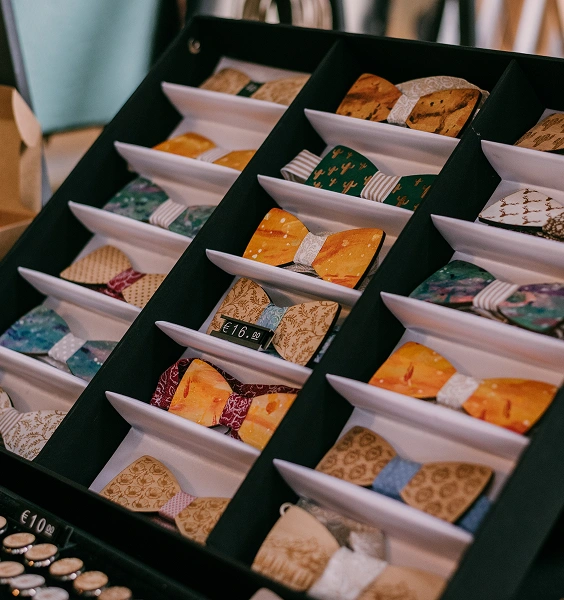 Na zdjęciu znajduje się stojak z różnorodnymi muszkami, poukładanymi w białych talerzykach. Muszki mają różne wzory i kolory, a cena każdej z nich jest widoczna obok.